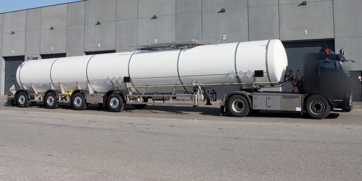 White chemical tank semi-trailer behind a tractor unit outside a tank builder's works, the equipment a chemical tanker carrier runs